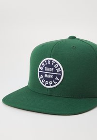 Green snapback cap made of textured fabric. Features a circular stitched patch with “Brixton Supply” in navy and white. Flat brim design.