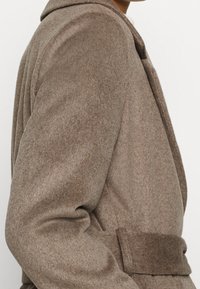 Bruine wollen mix jas met brede revers en een bijpassende stoffen riem, gedragen door een persoon die van schouder tot taille in profiel is afgebeeld.