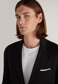 Schwarzer Blazer aus glattem Stoff, mit einer Brusttasche und einem weißen Taschentuch, kombiniert mit einem schlichten weißen T-Shirt darunter.