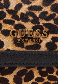 Sekk med leopardprint, der har en sort struktureret rem, et metalisk guld "GUESS EST 1981" logo og en pelslignende overflade.