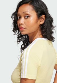Yellow fitted t-shirt with white three-stripe accents on shoulders, short sleeves, and a smooth texture. Model's hair is curly and dark.