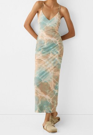 Mujer que lleva un vestido sin mangas, con escote en V, hasta el tobillo, con un patrón de teñido anudado en azul y beige y zapatos beige tipo mocasín con detalle de hebilla.