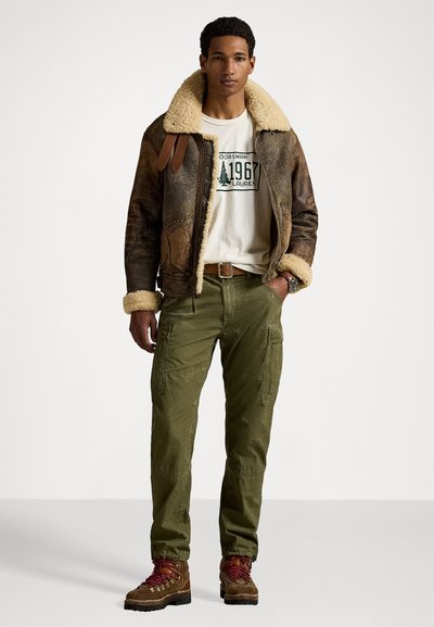 Bruine leren jas met schapenvachtvoering, crème t-shirt met groene print, olijfgroene cargobroek en bruine wandelschoenen met rode veters.