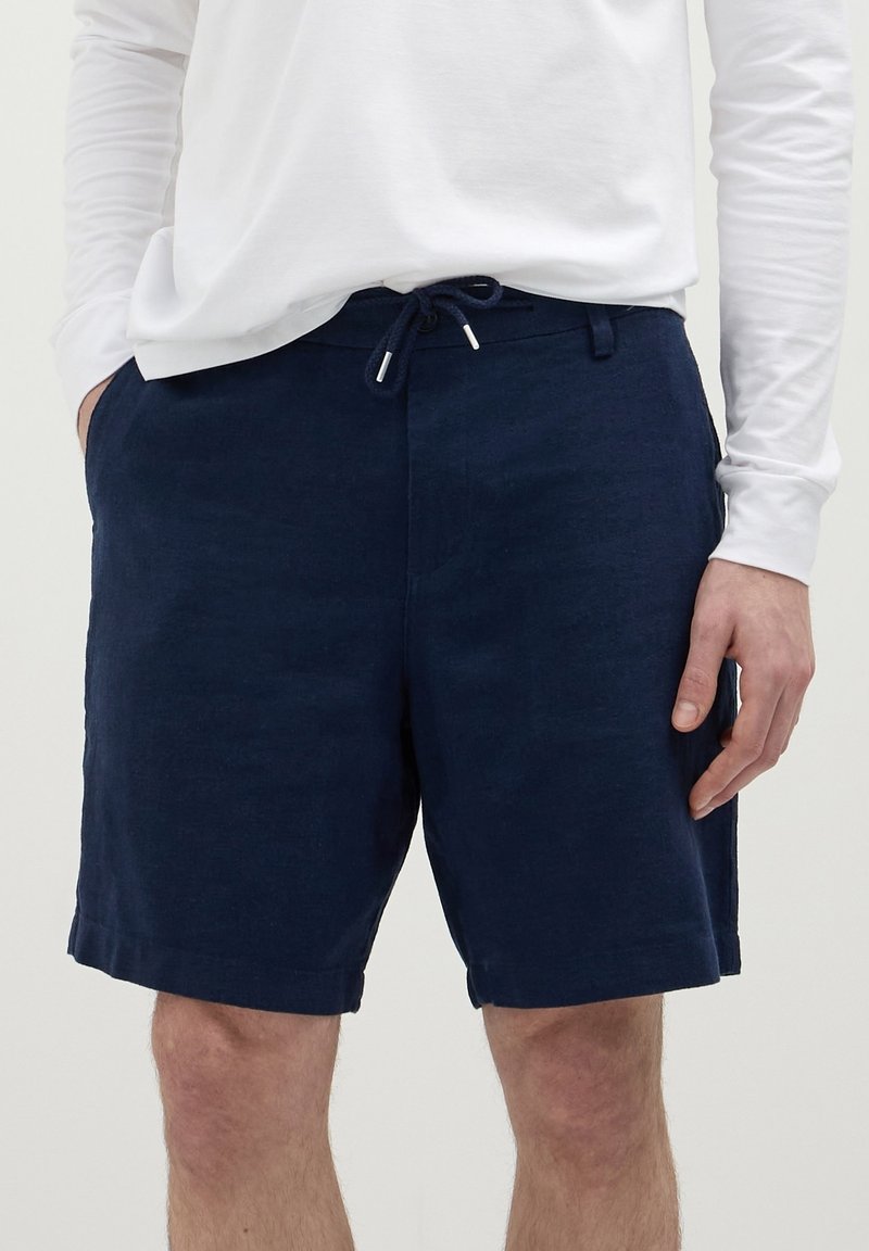 Homme portant un short bleu marine à cordon et une chemise blanche à manches longues, les mains dans les poches, debout devant un fond uni.
