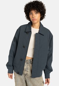 Young person with curly hair wearing a dark blue buttoned jacket over a beige cropped top and light-wash jeans against a plain background.