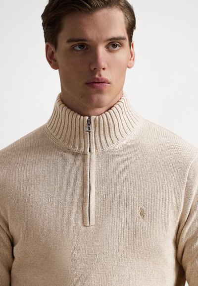 Beige gebreide trui met een hoge geribde kraag, voorzien van een rits tot een kwart van de lengte en een klein geborduurd logo op de borst.