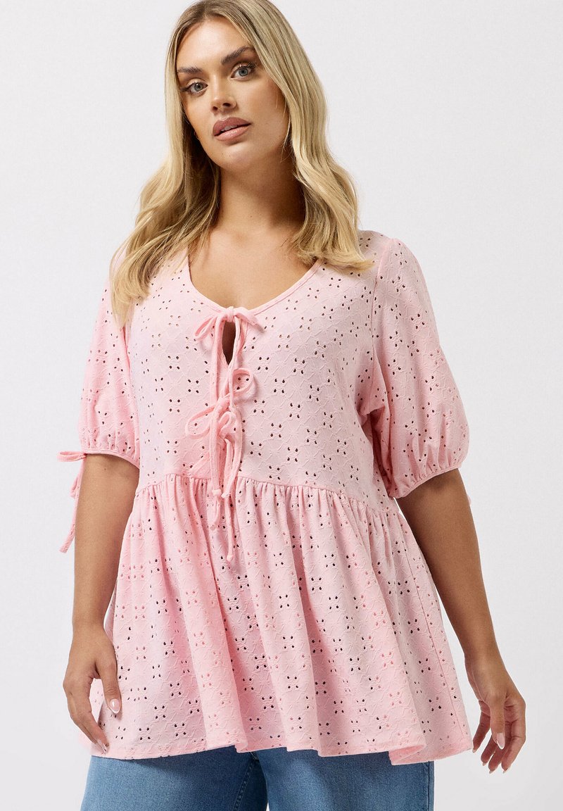 Blondynka w jasnoróżowej bluzce z ażurowej tkaniny z wiązaniem z przodu i bufkami o długości do łokcia, w połączeniu z niebieskimi dżinsami, na białym tle.