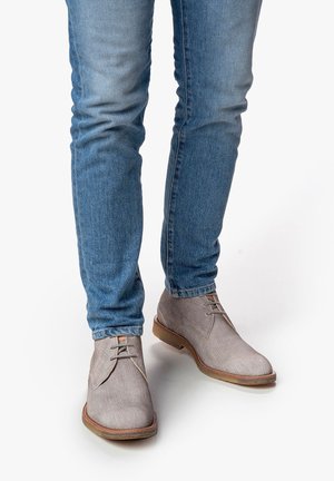 Hellgraue Stoff-Ankle Boots mit glatter Textur, Schnürdesign und kontrastierender brauner Sohle, kombiniert mit blauen Slim-Fit-Jeans.