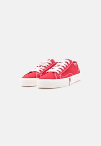 Ζευγάρι από χαμηλά πανί κόκκινα αθλητικά παπούτσια με λευκά κορδόνια και χοντρές λευκές λαστιχένιες σόλες σε απλό φωτεινό φόντο.