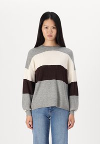 Pull en tricot avec des rayures grises, crème et bordeaux. Encolure ronde, manches longues, texture côtelée et coupe décontractée. Associé à un jean bleu clair.