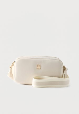 Cream rectangular crossbody bag with gold logo on front and wide matching adjustable strap coiled beside it.
