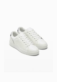 Sneaker bianchi realizzati in materiale sintetico liscio, con punta arrotondata, chiusura con lacci e suola in gomma texturizzata con un accento argento sul tallone.