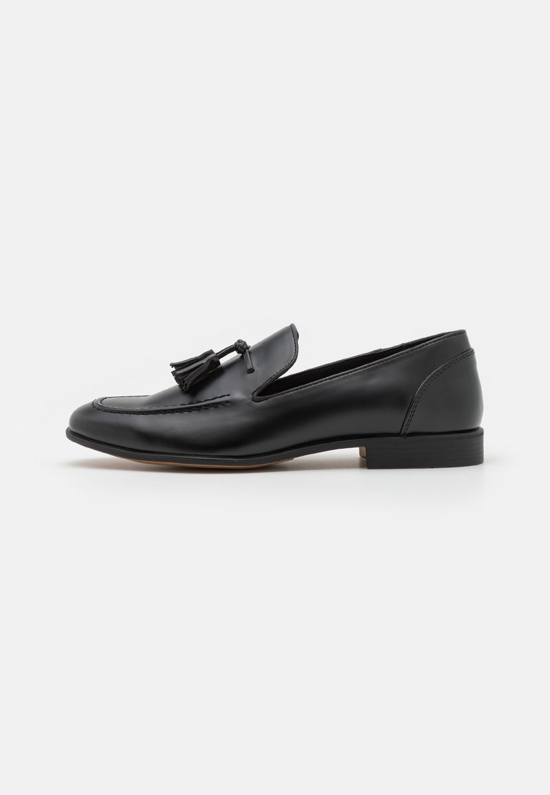 Mocassin en cuir noir avec pampilles, talon bas et détails cousus sur la tige, présenté en profil latéral sur fond blanc.