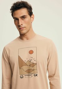 Man wearing beige long-sleeve shirt with graphic of brown hills, orange sun, flying birds, and text "HENDERSON" on light background.