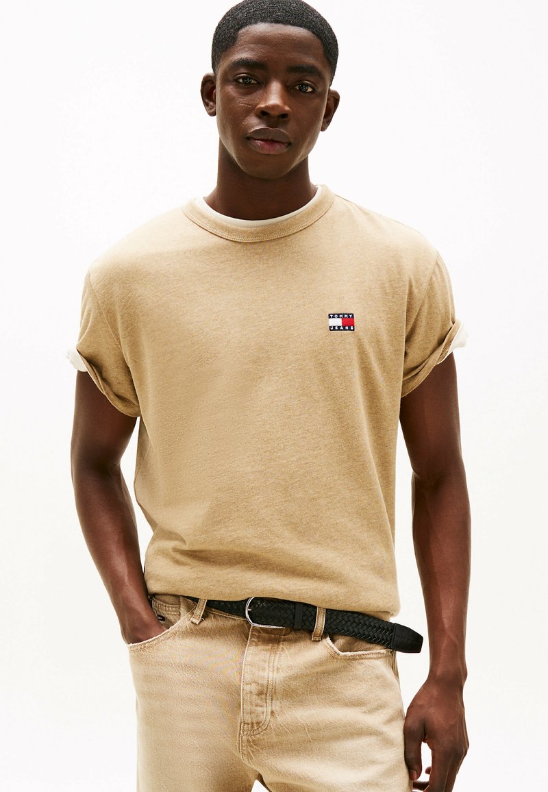 T-shirt beige in cotone con maniche arrotolate e una piccola patch con logo in rosso, bianco e blu; abbinato a jeans denim beige chiaro e una cintura intrecciata nera.