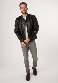 Giubbotto di pelle nera con zip frontale, colletto a costine e polsini. Indossato sopra una maglietta bianca, abbinato a jeans grigi e stivali marroni.