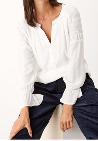 Camicia bianca con scollatura a bottoni e dettagli arricciati su spalle e maniche. Tessuto morbido con maniche fluide e voluminose.