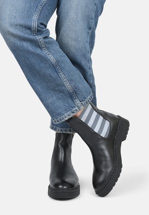 Sorte læder ankelstøvler med elastiske sidestykker og gummisål, parret med lysegrå og hvide stribede sokker, bæres med blå jeans.