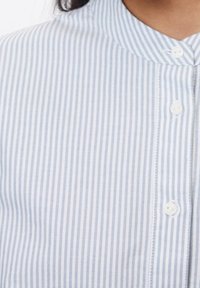 Ljusblå och vit randig skjorta med knappar och rundad krage, som har små vita knappar och en mjuk tygstruktur.