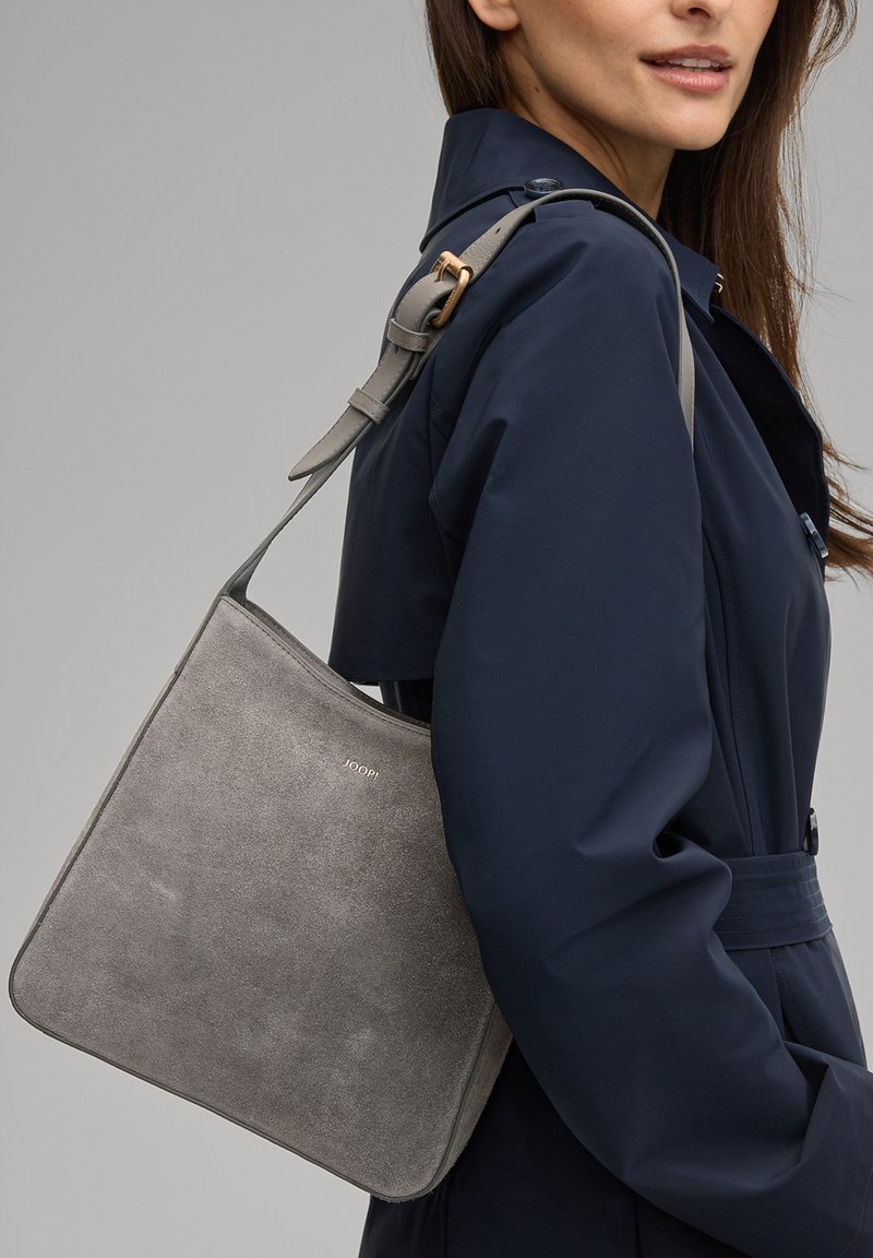 Mujer con abrigo largo azul marino que lleva un gran bolso de hombro de ante gris con correa ajustable y hebilla dorada.