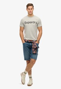 Grå t-shirt med sort logo, denimshorts, ternet skjorte knyttet om taljen, hvide sneakers med neutrale accenter, og stribede sokker.