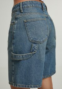 Gaiši zili džinsu šorti ar augstu jostasvietu, četriem kabatām un brīvu piegriezumu. Audumam ir teksturēta virsma ar redzamām šuvēm.