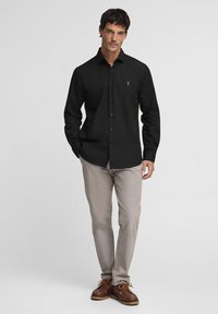 Camicia nera con bottoni e maniche lunghe, dotata di un logo discreto sul petto sinistro, abbinata a pantaloni beige chiaro e scarpe marroni.