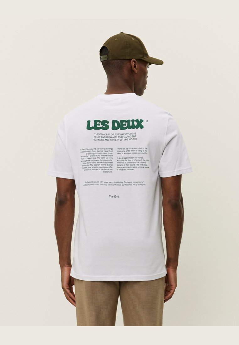 Biela bavlnená tričko s grafikou "LES DEUX" v zelenej farbe na chrbte, obsahujúca text o konceptoch a dizajne susedstva.