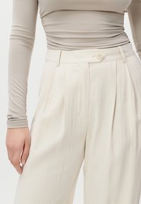 Langermet taupe topp med rynker, sammen med høyhåndte kremfargede bukser med folder, knappelukking og en glatt tekstur.
