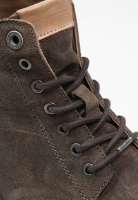 Stivaletto in suède marrone con superficie texturizzata, occhielli in metallo, lacci marroni e un accento in pelle beige sul colletto.