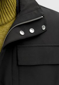 Čierna bunda s textúrovaným povrchom, vybavená zapínaním na zips, patentnými gombíkmi a kontrastným zeleným pleteným golierom.