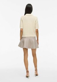 Crème gebreide cardigan met korte mouwen en kraag, gecombineerd met een beige uitlopende rok. Model draagt lichte sandalen met bandjes.
