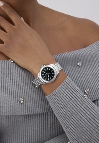 Orologio d'argento con quadrante nero, cinturino in metallo e indicatori luminosi, indossato su una mano. Maglione grigio a coste con decorazioni.