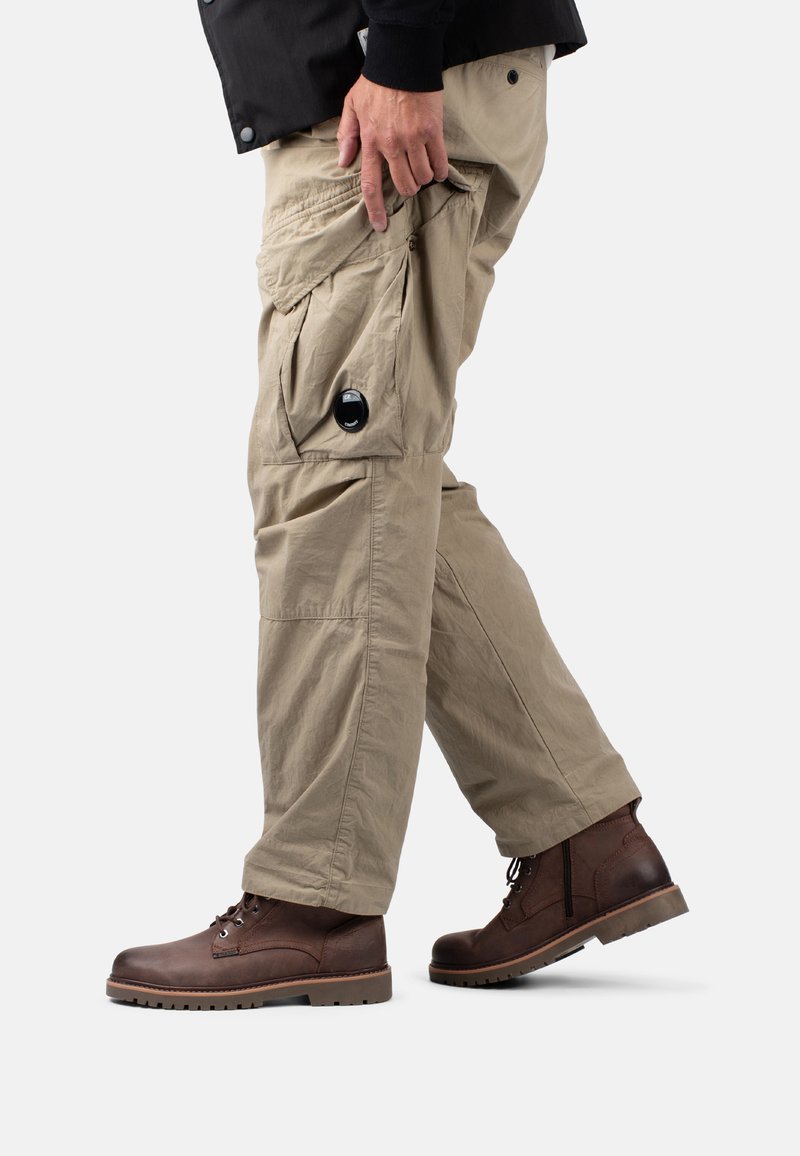 I pantaloni cargo beige presentano molteplici tasche e un dettaglio con bottone. Abbinati a stivali di pelle marrone, che mostrano un design robusto e una finitura testurizzata.
