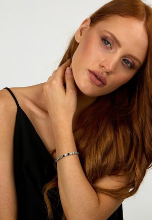 Silbernes Armband mit rechteckigen Gliedern und klaren Edelsteinen. Am Handgelenk getragen, ergänzt durch weiche, wellige rote Haare und ein schwarzes Oberteil.