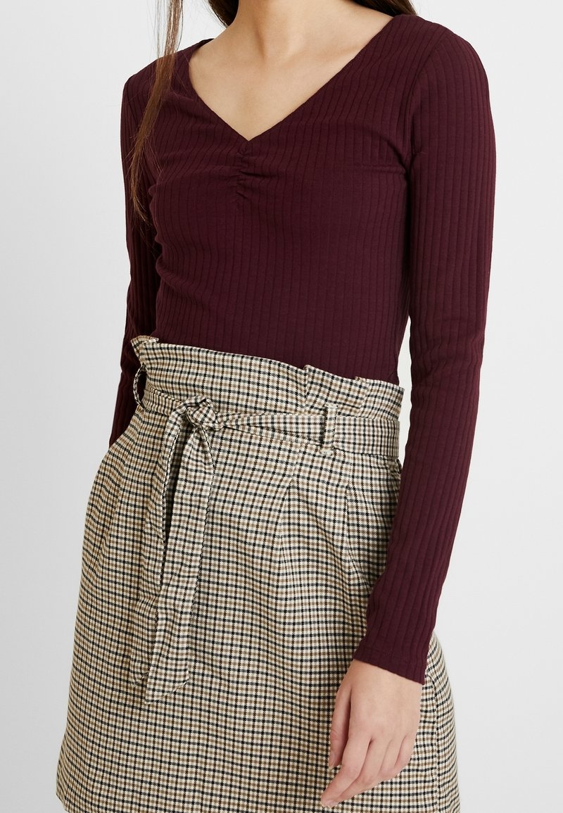 Žebrovaný vínově červený top s dlouhým rukávem a výstřihem do V, doplněný sukní s vysokým pasem a kostičkovaným vzorem, která má ozdobný pásek.