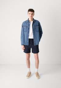 Camisa de mezclilla de botones con dos bolsillos en el pecho, camiseta blanca, pantalones cortos azul marino y zapatillas beige. El modelo está de pie sobre un fondo liso.