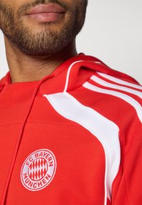 Rød hættetrøje med hvide detaljer, prydet med FC Bayern München-logo. Fremstillet af blødt stof med synlig tekstur og sporty designelementer.
