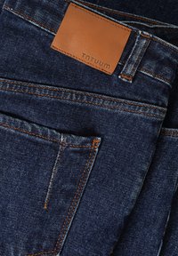 Jeans de mezclilla azul oscuro con un parche de cuero marrón que dice "TNTUUM", con costuras naranjas y un diseño clásico de cinco bolsillos.