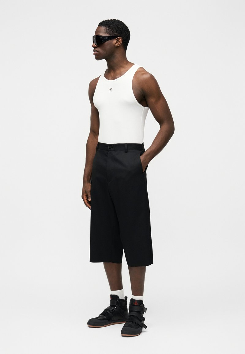 Débardeur blanc, short noir longueur genoux, bottes noires à lanières, et grandes lunettes de soleil. Design minimaliste et coupe décontractée.