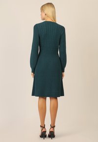 Teal strikket kjole med en tætsiddende kabelstrik overdel, lange ærmer og en udvidet nederdel. Har strukturerede mønstre og en glat overflade.