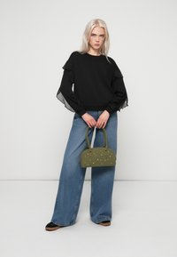 Czarny sweter z falbaniastymi, przezroczystymi rękawami, w połączeniu z niebieskimi dżinsami o wysokim stanie i szerokich nogawkach. Model trzyma małą, oliwkową torebkę ozdobioną gwiazdkami.