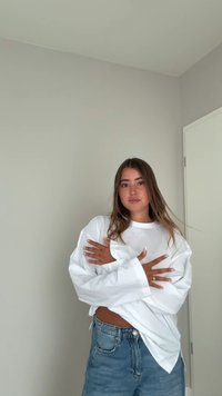 Überdimensioniertes weißes Langarmshirt mit herabfallenden Schultern, kombiniert mit hoch taillierten blauen Jeanshorts. Glattes Material, entspannte Passform.
