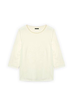 Kermanvärinen puuvillapaita, jossa on kolme neljäsosaa hihat, keskiosassa teksturoitu kukkakuvio ja koristeelliset yksityiskohdat hihoissa.