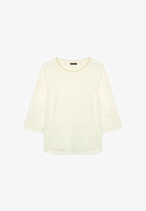 Cremfarbendes Baumwolloberteil mit drei Viertelärmeln, das in der Mitte ein strukturiertes Blumenmuster und dekorative Akzente an den Ärmeln aufweist.