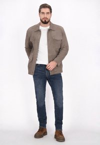 Taupe Wildlederjacke mit vier Taschen, Knopfverschluss, kombiniert mit einem weißen T-Shirt und dunkelblauen Jeans. Braune Schnürschuhe.