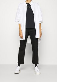 Witte oversized button-up shirt over een zwart t-shirt, gecombineerd met zwarte broek en witte sneakers. De outfit heeft een ontspannen pasvorm.