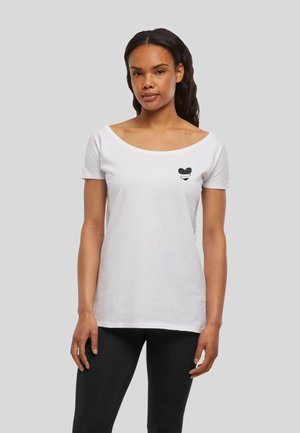 Femme aux longs cheveux foncés portant un t-shirt blanc à col rond avec un petit motif cœur noir et un pantalon noir, debout devant un fond gris.