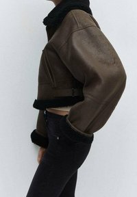 Ruskea nahkainen lyhyt takki, jossa on musta lampaanvilla-kaulus ja hihat, sivuvetoketju ja säädettävä vyö yksityiskohtana vyötäröllä.