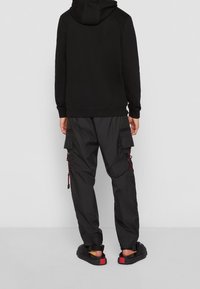 Felpa nera con cappuccio dal taglio rilassato, abbinata a pantaloni cargo neri con tasche laterali e dettagli rossi per i cordini. Includendo sandali neri.